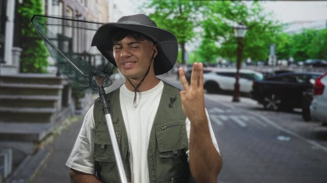 Man fisherman wearing a wide hat and vest holding a net and showing two fingers with his hand on a street in daylight; playful confidence.