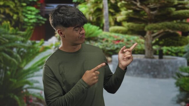 Man in olive long sleeve shirt points index fingers to the right with a wry smirk in a manicured garden courtyard; playful curiosity.
