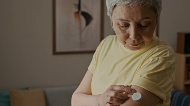 Tilt down shot of senior woman adjusting adhesive patch of CGM on upper arm while using medical technology in daily life at home, slow motion