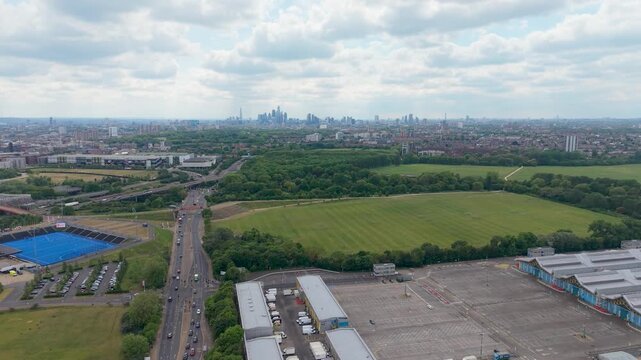 London skyline in distance as drone reverses over Hackney Marshes, New Spitalfields Market, Eastway and the A12 corridor. Wide aerial view of green fields, roads and city.
