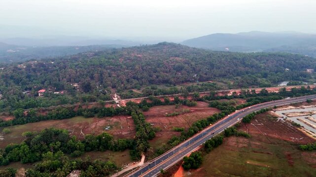 Drone shot capturing a scenic highway cutting across greenery and red earth terrain, surrounded by palm-covered settlements, coastal inlets, and distant hills under soft atmospheric light.