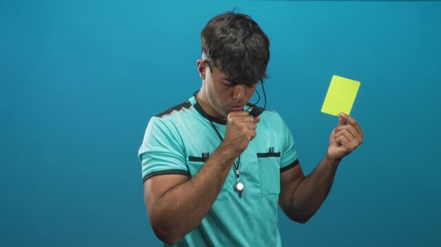 Man referee in studio holds yellow card, coughs into fist and displays whistle on lanyard; authority serious.