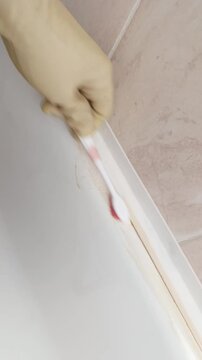 Gloved hand scrubbing bathtub seam vigorously using small toothbrush to lift stubborn brown stains and soap scum from silicone junction closeup of repetitive motion precise