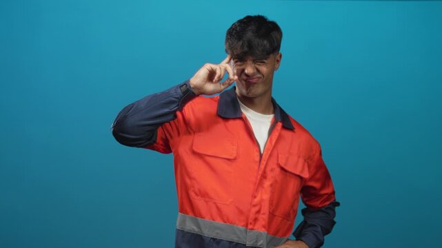 Man in orange work uniform points finger to temple and makes a smirk in studio against a blue backdrop; playful gesture.