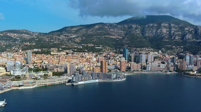 4K aerial view of Monaco from the sea with Mareterra development, Tour Od&eacute;on skyscraper, dense residential towers, Larvotto beach and Mont Agel mountain backdrop under moody clouds.