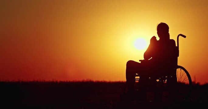 Silhouette of a disabled woman sitting in wheelchair with her hands together praying, background of a golden sunset.