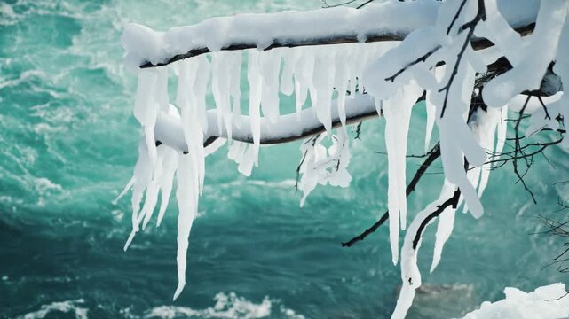 Close up view of snow-covered branches and icicles dangling above a vibrant blue river, dynamic winter scenery with flowing water.