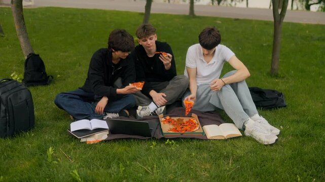 Wide shot of three teen male students taking pizza slices on park blanket