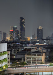 Obraz premium A cluster of gleaming skyscrapers pierces the heavy twilight sky, casting a brilliant array of golden and white lights over the expansive cityscape below. In the foreground, a tapestry of low-rise nei