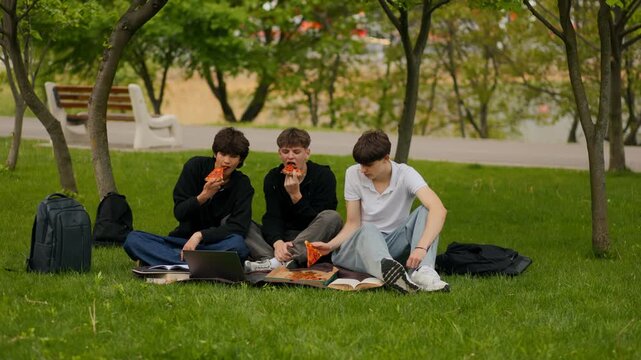 Wide shot of three teen male students sharing pizza slices on grass