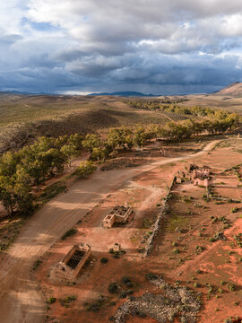 Aerial view of the historic Kanyaka Station ruins and a winding dirt road surrounded by arid landscape and trees under a cloudy sky in Flinders Ranges, South Australia, Australia.
