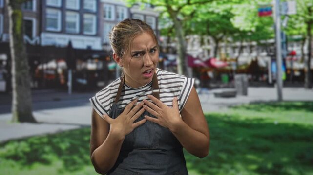 Woman in apron with hands on chest and puzzled expression standing on a street near outdoor cafes and tables; concern.