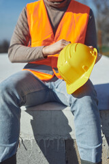 Fototapeta premium Construction worker working outdoors on a construction site, making a pause and holding a yellow hardhat.