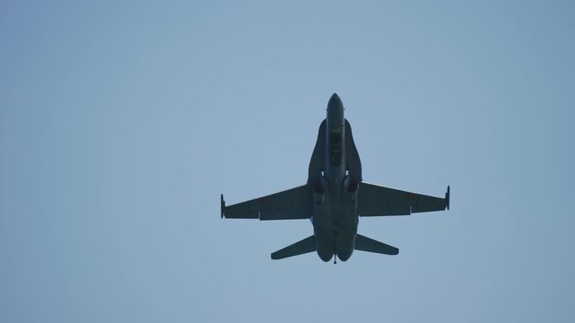 Military fighter jet flies directly overhead against clear blue sky. Camera follows aircraft as it gets closer, revealing details of its undercarriage and wings. Powerful and dynamic shot