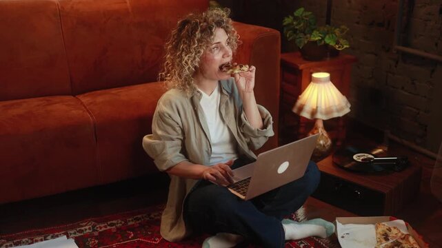 Curly haired woman eating pizza while working remotely on laptop and taking break during online work at home. High quality 4k footage