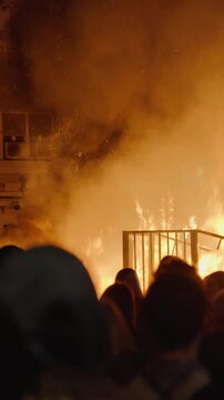 Large flower sculpture on burning falla pole during Las Fallas crema in Valencia, Spain, black smoke billowing over building as crowd watches from street