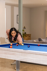 Obraz premium Asian woman aiming pool shot on blue-felt table with cue-stick at home in pink sweater