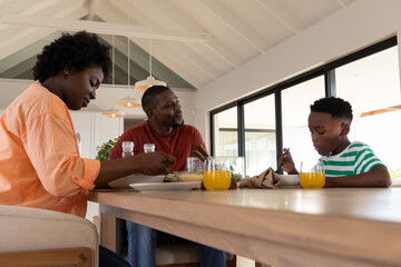 African American family sitting at wooden dining table inside modern home, sharing orange juice © wavebreak3