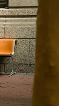 Vertical video: Entering, woman in yellow coat walking on sidewalk, blocking orange bench, bike
