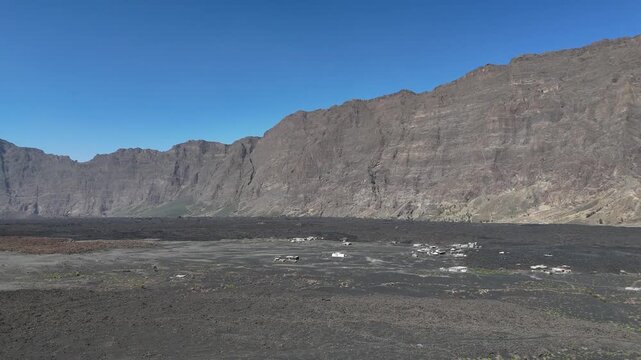 Wide drone shot of the Ch&atilde; das Caldeiras volcanic plain and the massive Bordeira cliff wall in Fogo National Park, Cabo Verde, featuring remote village settlements on black lava soil