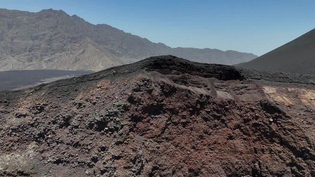 Close-up drone shot of the volcanic crater rim of Pico do Fogo, showing the dark, porous basaltic rocks and the vast Ch&atilde; das Caldeiras plain with the Bordeira wall in the distance