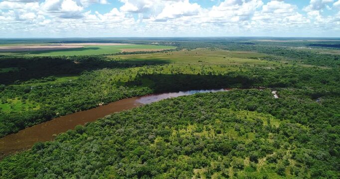 Meandering cerrado river flowing beside wet grazing land, gallery forest and agricultural frontier near Arinos, Minas Gerais, Brazil in the Urucuia Plateau region. Drone forward flyover.