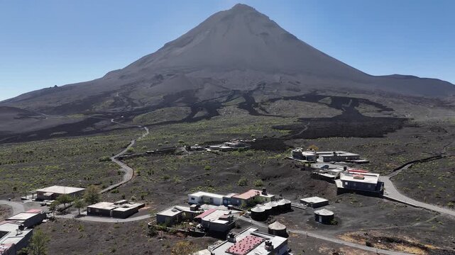 Aerial wide shot of the majestic Pico do Fogo volcanic cone towering over the small settlement of Ch&atilde; das Caldeiras, featuring winding roads and expansive dark lava fields in Cabo Verde
