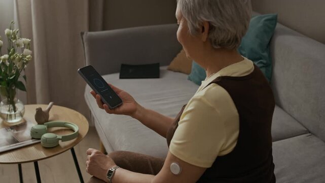 Over shoulder shot of senior woman with diabetes CGM device on arm holding smartphone while recording voice command to AI assistant in living room, dolly in camera movement