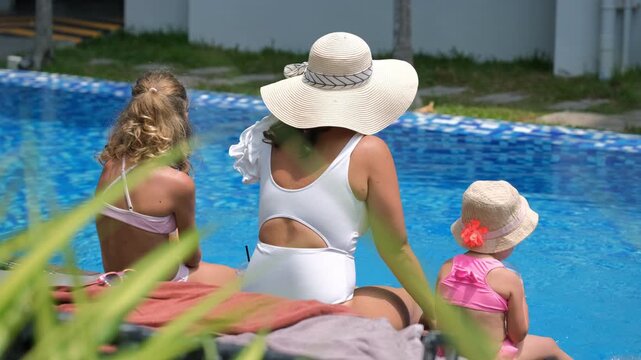 Rear view of young mother and daughters sitting at pool edge dangling feet in the water while little daughter sips a cocktail. Family holiday concept