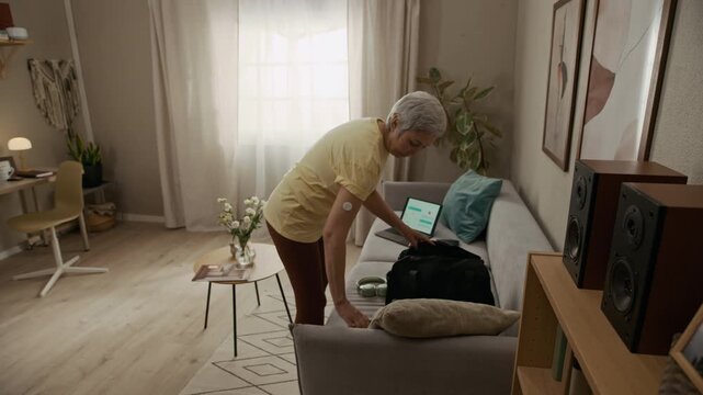 Wide angle shot of senior woman with diabetes wearing continuous glucose monitoring sensor on arm packing workout clothes into bag in living room before heading to gym