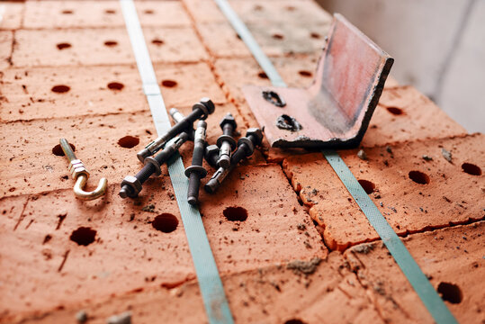 Metal anchor bolts, a threaded hook, and a steel angle bracket sit on top of a strapped stack of perforated red clay bricks at a construction site.
