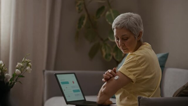 Side view medium shot of senior woman pausing from remote work on laptop to check adhesive patch on upper arm while managing diabetes with CGM device at home