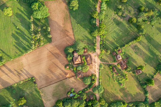 Aerial view of ancient Buddhist temples and pagodas scattered across lush green fields and dirt paths under warm sunlight in Old Bagan, Mandalay Region, Myanmar Burma.