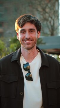 A cheerful young man wearing stylish sunglasses and a casual outfit stands outdoors, smiling warmly in a vibrant setting illuminated by the golden hour light, creating a delightful atmosphere.