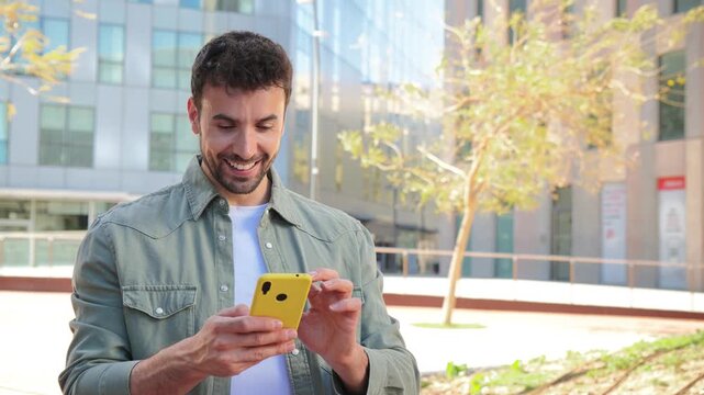 Cheerful man happily using smartphone, immersed in an enjoyable interaction, showcasing positive emotions and engagement with technology in a lively outdoor space.