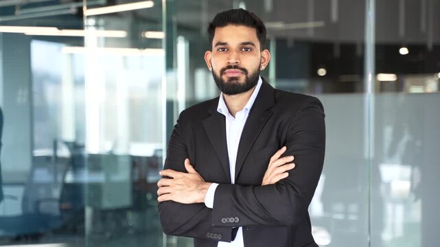 Portrait of serious businessman in formal suit standing with crossed arms at workplace in a modern glass business office. Confident successful handsome manager or worker posing looking at camera