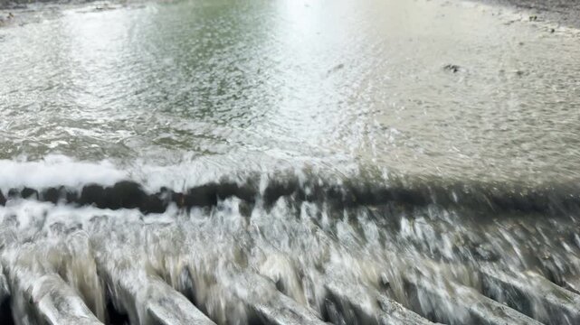 Turbid rain surges over a concrete edge and into a metal grate. This fast motion creates a loud, white-noise roar and a constant splashing sound.