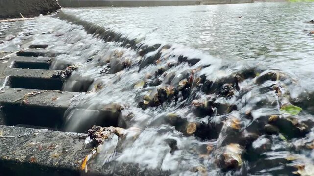 Heavy rainwater surges over a concrete edge and into a metal grate. This rapid movement produces a loud, churning roar and sharp splashing sounds.