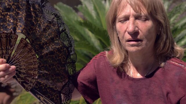 Mature blonde woman uses a large hand fan to cool herself during an extremely hot summer day while standing outdoors in a bright garden under strong sunlight