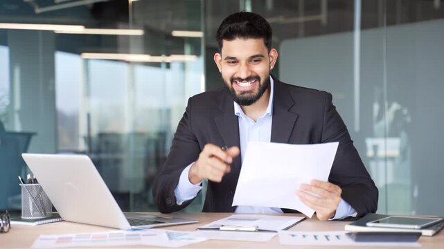 Happy businessman is satisfied with results of financial report checking documents sitting in business office. Manager in suit celebrating success, happy with positive indicators and looking in camera