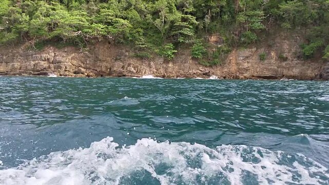 Dynamic boat wake churns azure tropical waters, revealing a scenic journey towards a rugged coastline of vibrant green cliffs and dense forest