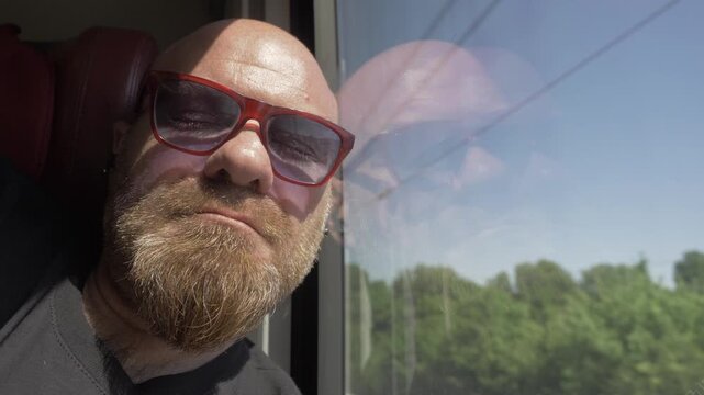 Extreme close-up of a bald man with a red goatee sleeping peacefully against the train window during a bright daytime journey.