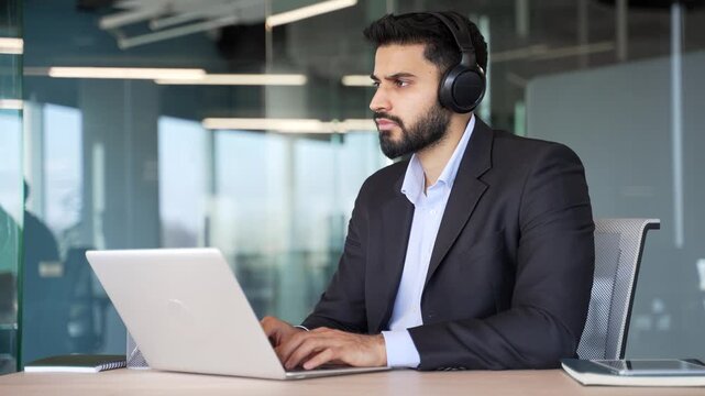 Focused businessman in formal suit and headphones works on laptop sitting at a workplace in a business office. Serious coder developer or IT specialist is programming, busy with a project on computer