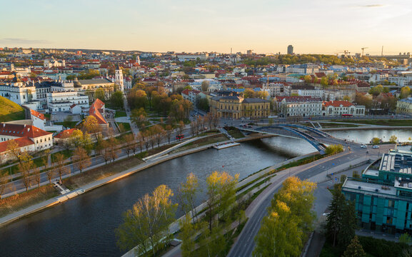 Aerial view of the Vilnius city skyline and Neris River featuring the Mindaugas Bridge, historic architecture, and lush green trees during a golden sunset Vilnius County, Lithuania.