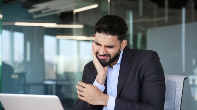 Upset businessman suffering from toothache while sitting at workplace in business office. Male worker in formal suit holds his hand on his cheek, massaging sore spot. He needs to see dentist. Close up