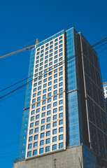 Obraz premium Modern glass skyscraper under construction with a yellow tower crane on top against a clear blue sky. Low angle view of contemporary urban development and high-rise architecture in Kyiv, Ukraine.