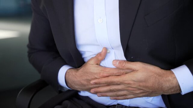Close up. Businessman in formal suit feels stomach pain sitting at workplace in business office. Male hands holding massaging stomach. Sick man worker suffers from gastritis, poisoning or bloating