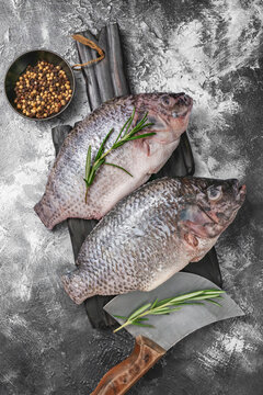 Two fresh raw tilapia fishes on a dark wooden board with rosemary and peppermorns.