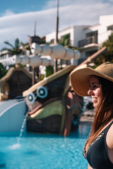 Woman smiles near water park slide. She wears sun hat and swimsuit. Palm trees and buildings frame background. Playful pirate ship shape adds fun vibe. Ideal for vacation, summer, or leisure themes © AlbertoRodriguez
