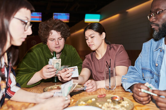 Multiethnic young adults counting cash and splitting restaurant bill at table, discussing shared expenses and budgeting. Useful for personal finance, social spending, cost sharing